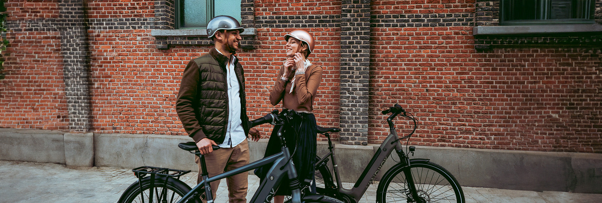 Deux cyclistes portant des casques de vélo Thousand en coloris polished titanium et rose gold, posant avec leurs vélos devant un mur de briques.